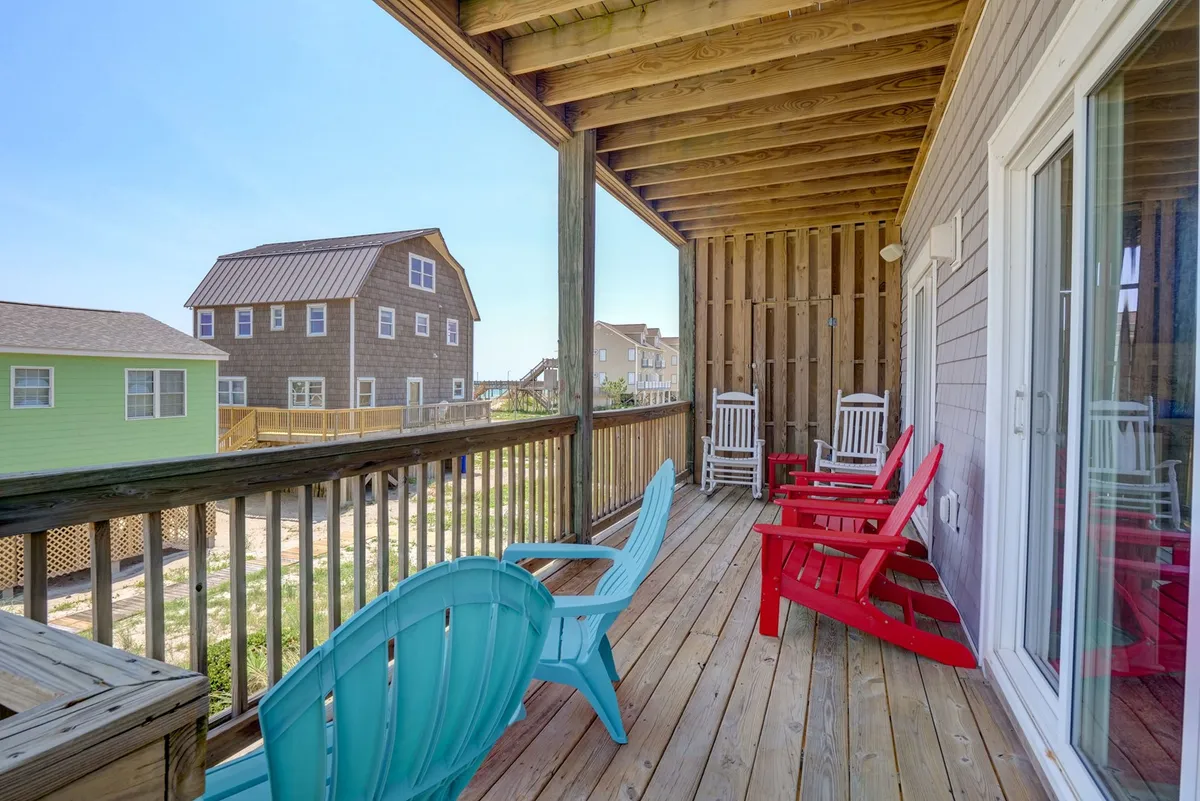 A spacious wooden deck overlooking the coastline, furnished with colorful Adirondack chairs and bordered by a simple railing. In the distance, neighboring houses hint at the duplex setting and a lively, communal atmosphere. The deck is sunlit, inviting guests to gather for coffee, group meals, or sunset relaxation while enjoying ocean breezes.