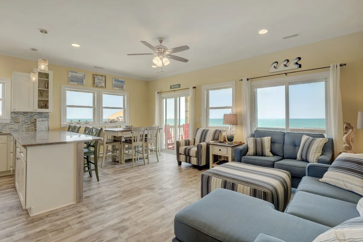 A comfortable open-concept living and dining area is shown, featuring a cozy sectional sofa, large windows with sweeping views of the ocean, a dining area with bar stools, and decorative beach-themed accents. Natural light floods the space, and the layout encourages gathering and relaxation.