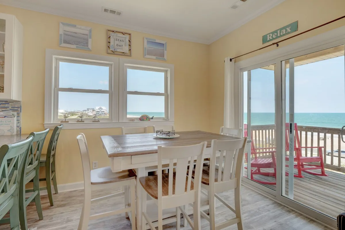 A bright and cheerful dining area is featured with a table for five, colorful chairs, and large windows framing beautiful ocean views. A 'Relax' sign and coastal touches add to the inviting atmosphere, making it ideal for meals or casual gatherings.