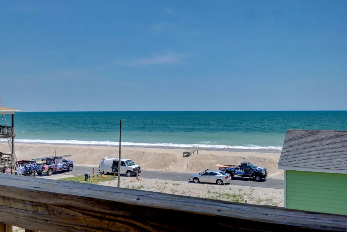 An expansive, sunlit ocean view with a sandy beach, blue sky, and the road nearby. The visual focus on the water, sky, and shoreline evokes a sense of openness and direct proximity to the coast, inviting guests to imagine easy days by the sea.