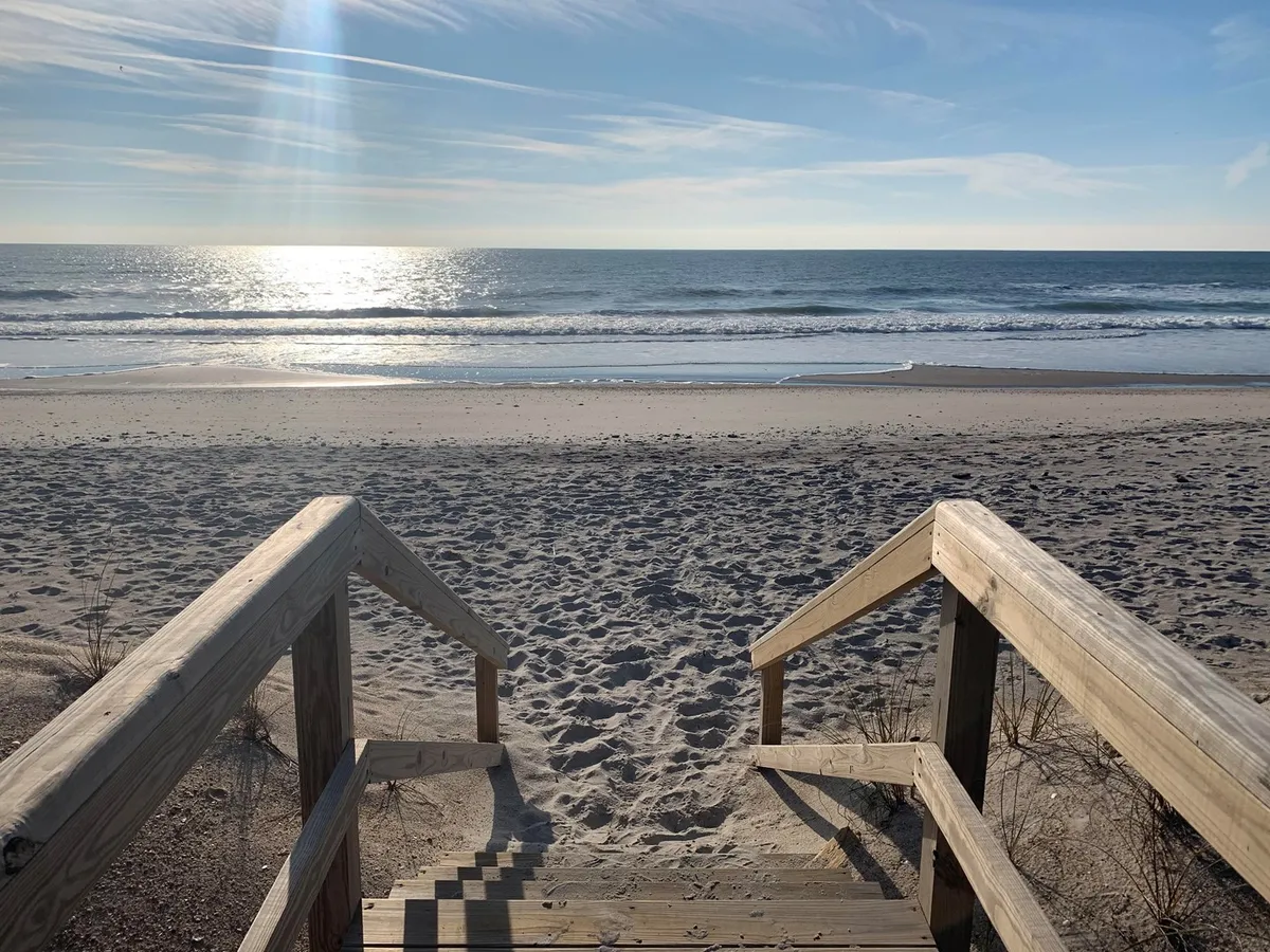 Wooden steps lead directly onto a serene, sandy beach with gentle sunlight sparkling on the water. The view highlights the close proximity to the ocean and the inviting, natural beauty of the coastline, ideal for romantic walks or quiet reflection.