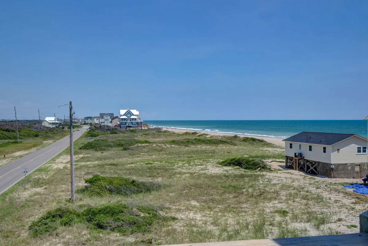 A panoramic view of the oceanfront with sandy shores, distant beach houses, and a quiet road nearby. The expanse of sand and water invites walks, picnics, and exploration, while the setting sun hints at ideal photo moments.
