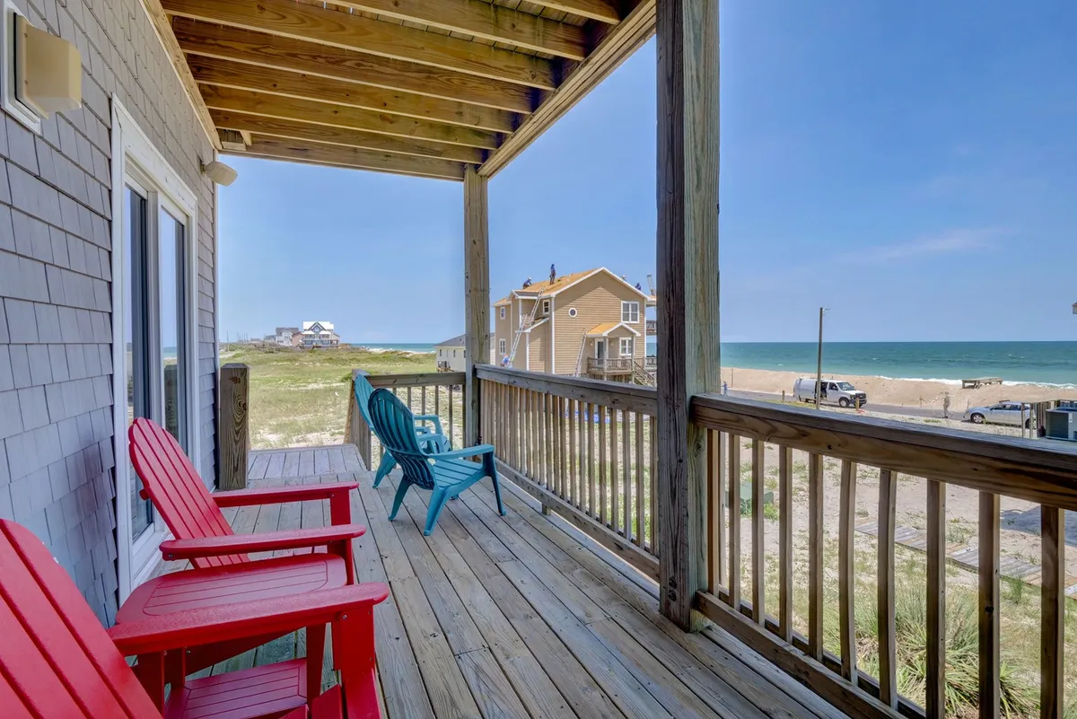 The hero image showcases a sun-kissed wooden deck lined with vibrant Adirondack chairs, all facing the endless blue of the Atlantic Ocean. The scene radiates tranquility and casual comfort, inviting guests to relax, sip coffee, or gather with friends while soaking in panoramic seaside views.