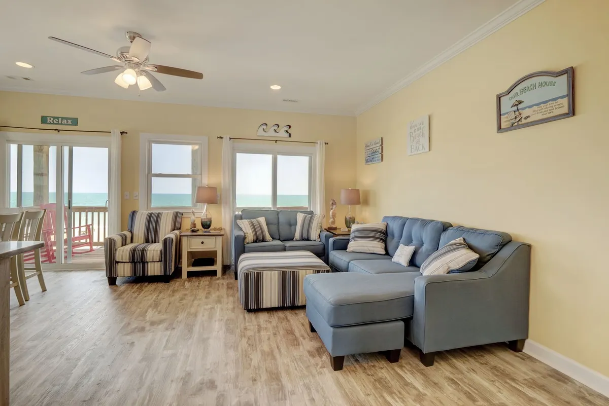 The living room is arranged with a large sectional sofa, a striped armchair, and ocean-facing windows. Decorative wall art and soft lighting create an inviting ambiance, and the layout encourages group relaxation or movie nights.