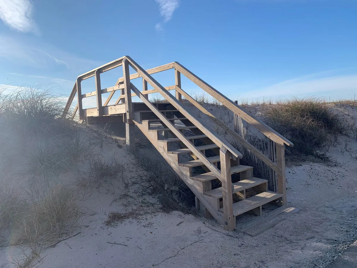 A sandy pathway leading from the wooden deck and staircase through dune grass to the beach. The inviting natural transition from home to beach scene encourages exploration and outdoor activity, perfectly matching the mention of paddleboarding, volleyball, and surf lessons.