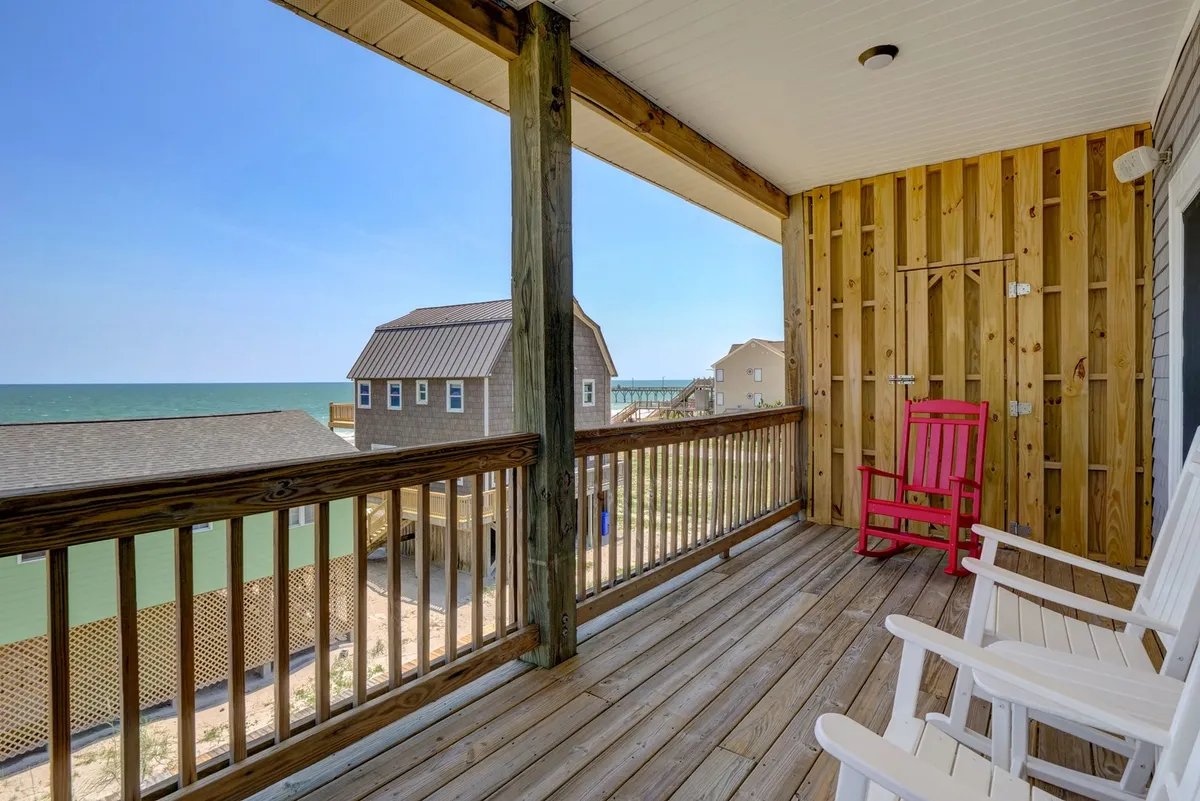 An oceanfront wooden deck with a row of colorful chairs facing the water. The expanse of blue ocean and clear sky create an inviting backdrop for group relaxation, conversation, and soaking in the views. The setting is ideal for evening gatherings or unwinding after a day of beach activities.