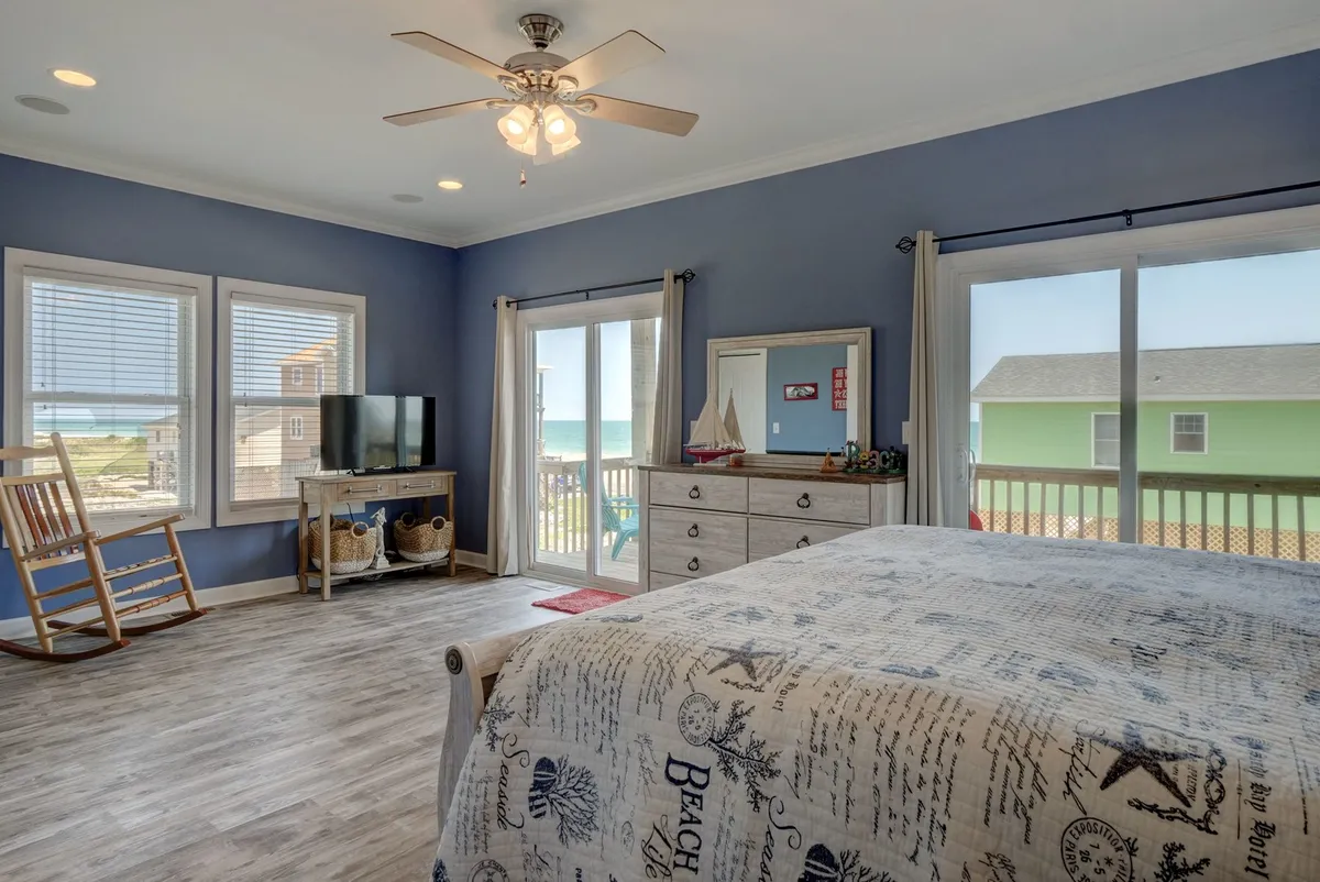 This image showcases a serene bedroom with nautical-themed bedding, large windows offering sweeping ocean views, a wooden rocking chair, and a dresser adorned with decorative items. The space is bathed in natural light, creating a peaceful atmosphere ideal for relaxation.
