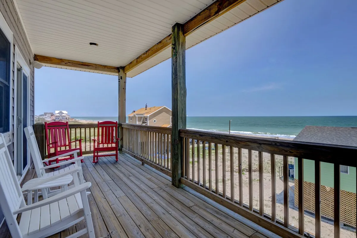 A tranquil exterior scene featuring two red rocking chairs positioned on a wooden deck, overlooking the ocean. The deck is framed by a wooden railing, and the view extends out to the open water with gentle waves and a soft, serene sky, evoking a sense of peace and quiet connection to nature.